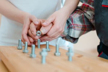 Detailaufnahme der Hände von zwei Menschen. Ein Mensch hilft dem anderen beim Zusammenführen von Muttern und Schrauben als motorische Übung