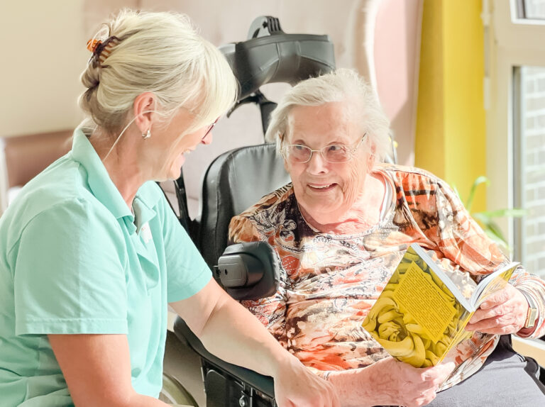 Eine Seniorin hält ein Buch in der Hand und unterhält sich mit einer Pflegerin