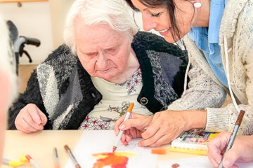 Eine Betreuungskraft hilft einer Seniorin beim Malen mit Filzstiften